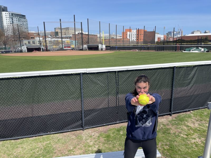 Girl holding up bright yellow softball with softball field in background.