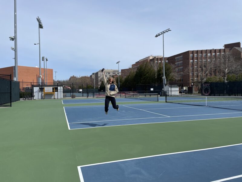 Girl jumping on a tennis court and smiling.