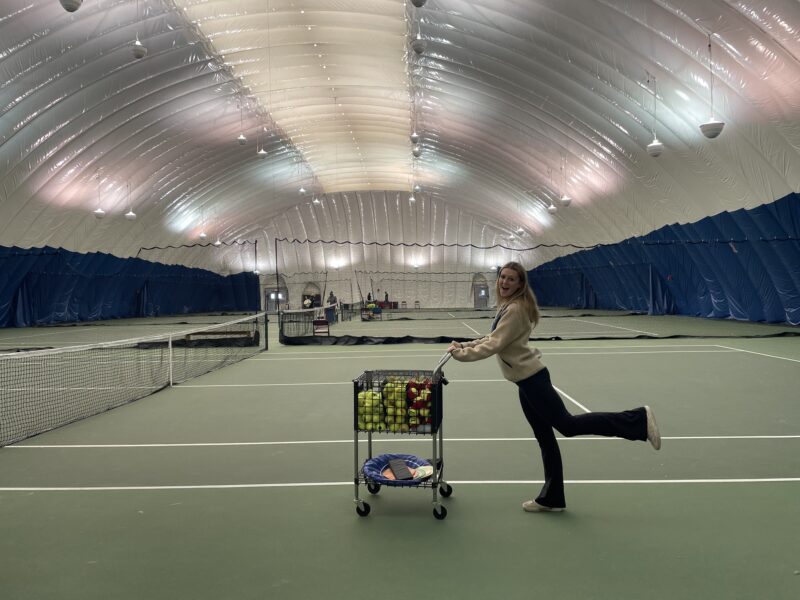 Girl pushing tennis ball cart inside an indoor tennis bubble.