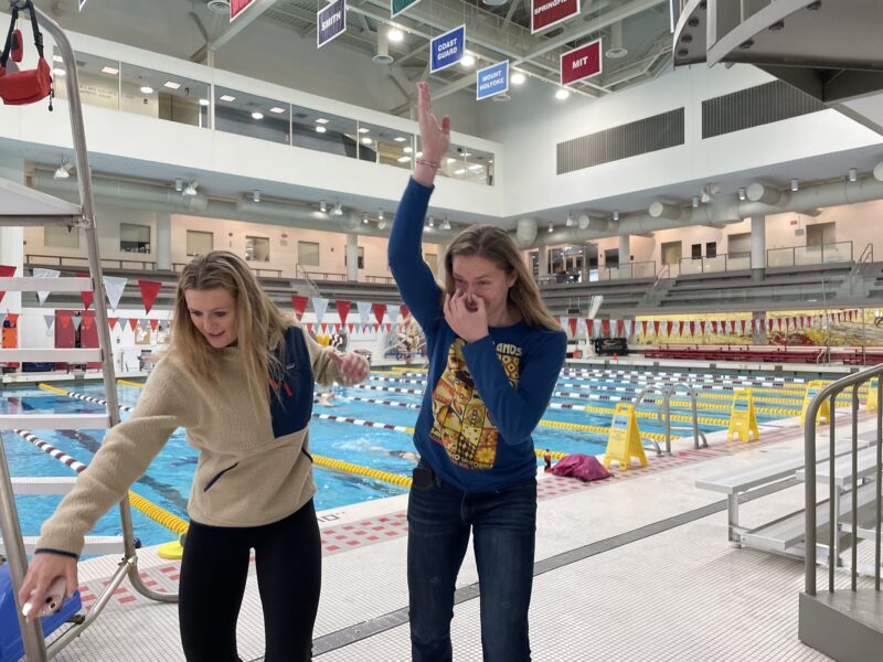 Two girls pretending to swim on land with swimming pool behind them.
