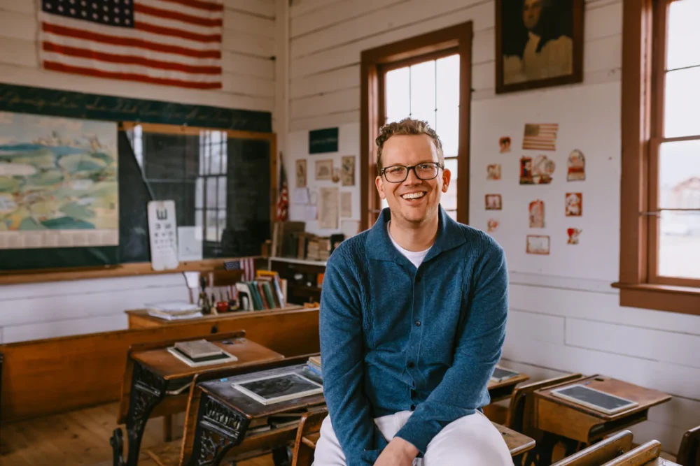 Hank Green sits smiling on an old wooden desk in a vintage classroom filled with books, posters, and a large American flag. He’s wearing glasses, a blue sweater, and white pants, with soft sunlight streaming in from the windows.