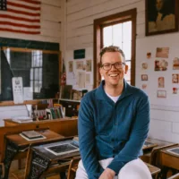 Hank Green sits smiling on an old wooden desk in a vintage classroom filled with…