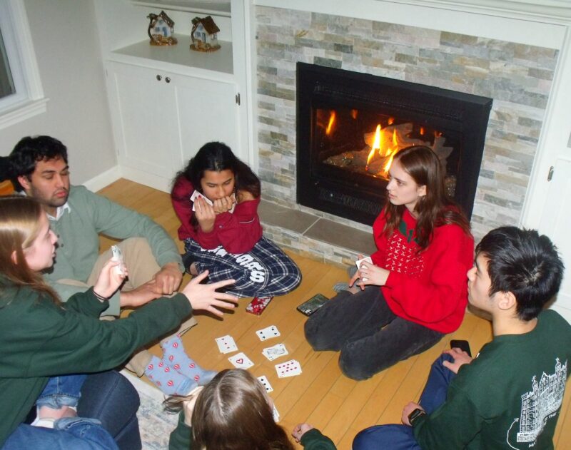 Six threeks sit cross-legged on a hardwood floor around a scattered circle of playing cards, facing a stone-trimmed gas fireplace with lively orange flames. They wear cozy sweatshirts and pajamas, some studying their hands, others gesturing animatedly. The mood is relaxed and warm.