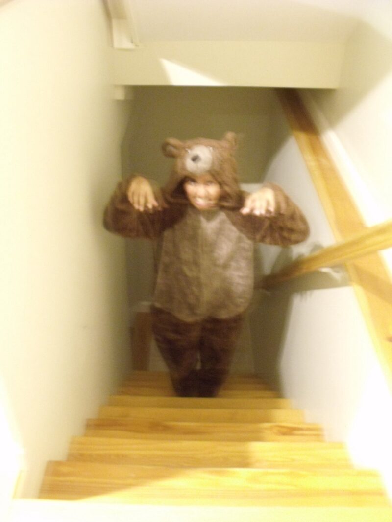 A blurry photo taken from the top of a staircase of a girl in a bear onesie. She is standing near the bottom of the stairs, hands mimicking claws and teeth mockingly bared
