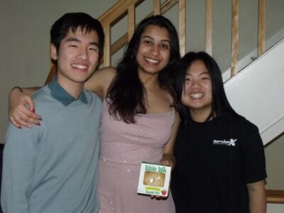 A boy and two girls standing together and smiling. The girl in the middle is holding a small apple pie in a box