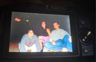 A photo of a camera displaying a picture of three young adults. The girl in the middle is smiling, the boys around her are staring into the camera