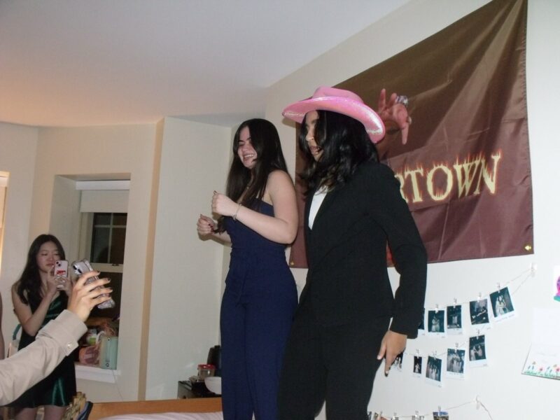 Two women, Anooshka and Emily, dance joyfully on a bed beneath the same “FLAVORTOWN” banner, Anooshka wearing a pink cowboy hat and black suit and Emily wearing a navy dress, laughing with her arms raised. Friends nearby snap photos and cheer them on. The wall behind them is decorated with rows of Polaroid photos. The vibe is celebratory and chaotic.