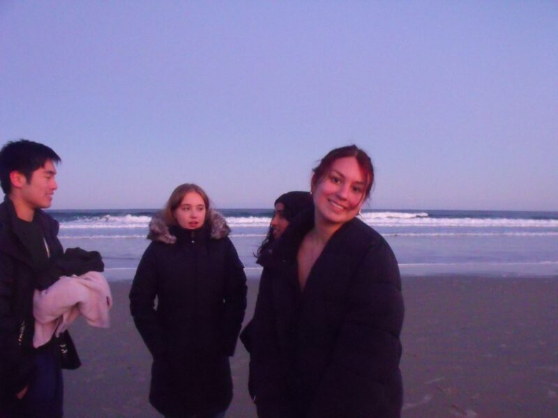 Four threeks stand on the beach, awash in the purple haze of the sunset. Three are mid conversation, while one of them is turned away from it smiling at the camera