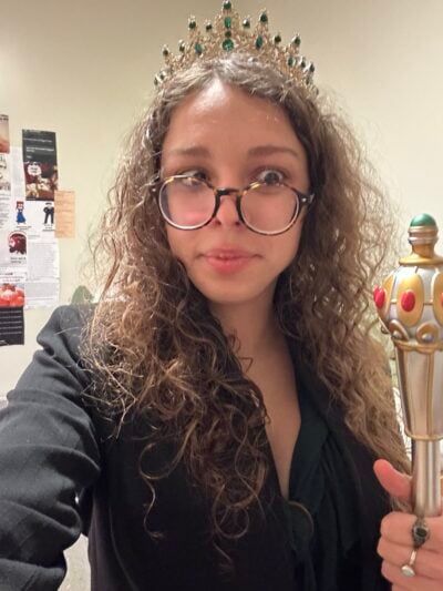 A selfie of a girl with semi curly brown hair, lopsided glasses, and a black blazer atop a green blouse holding a plastic royal staff in her left hand, which has a bronze and a silver ring on it. She is wearing a fake gold grown with green gemstones on it. Her brown glasses are lopsided and she has a wide-eyed, amused expression on her face