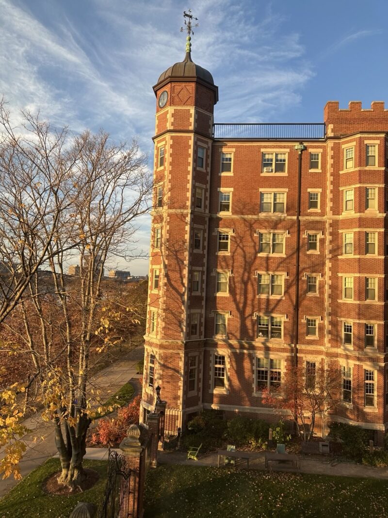 A photo of a castle-like brick dorm building, covered in windows. The walls are awash in a golden light from the sunrise