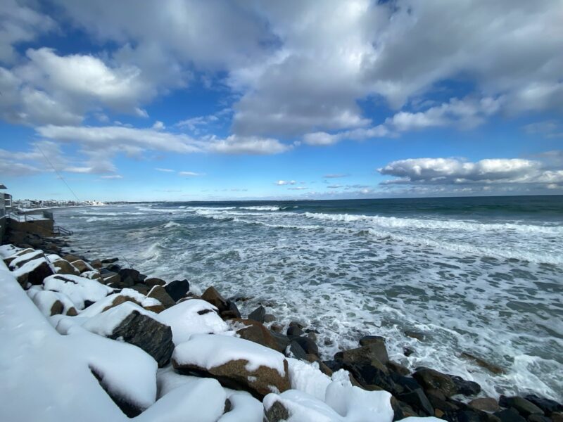A beach with a snow-covered rocky shore. The sea is foamy with waves, and the sky is partly cloudy but overall bright