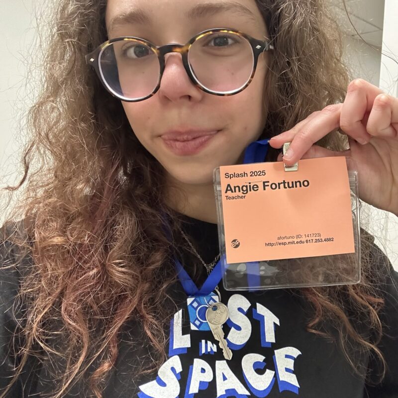 A girl with curly brown and red hair and glasses holds up a name tag reading