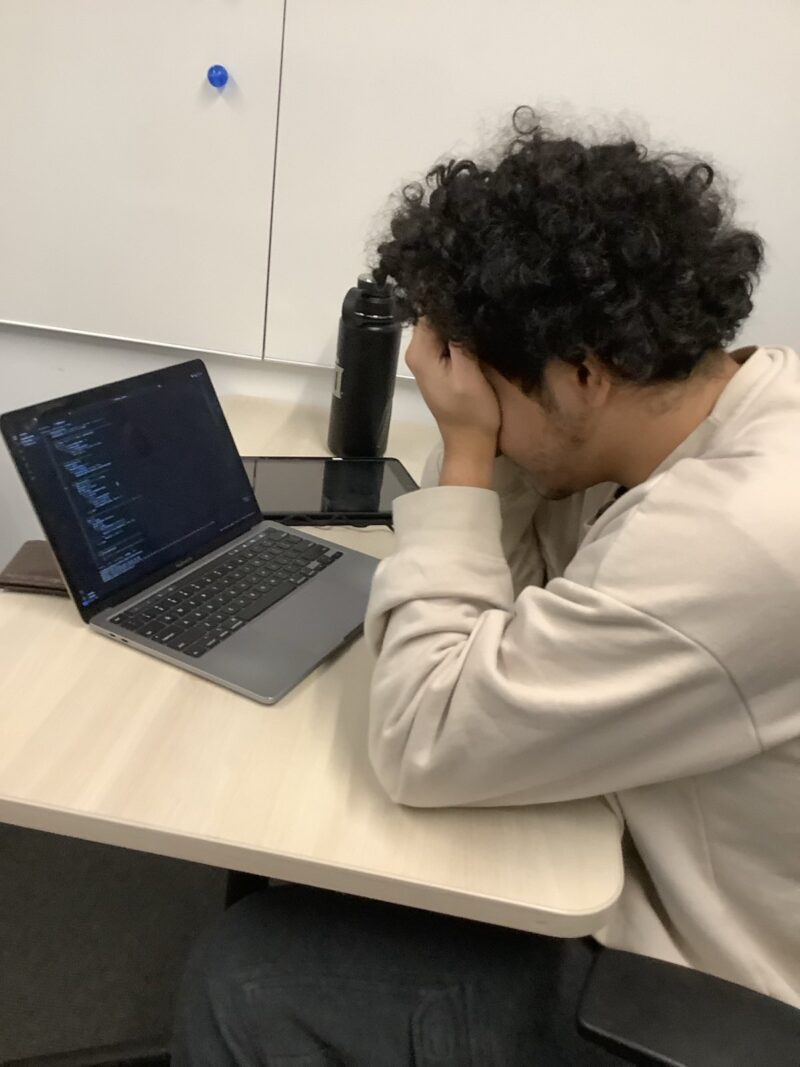 A picture of a boy with curly brown hair holding his head in his hands, a laptop with code open in front of him