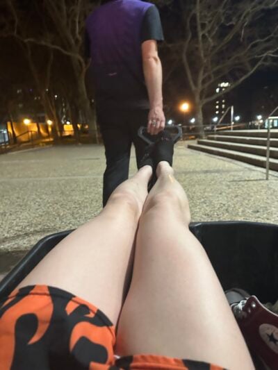 A picture taken from inside a wheelbarrow. Only my legs are visible. I'm wearing shirts with flame print and my right ankle is swollen. A man in a purple short is pulling the wheelbarrow through MIT campus at night.