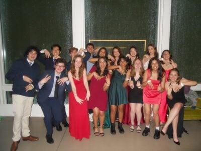 18 of the threeks, the MIT freshmen this post centers around, pose for a playful photo at our freshman formal, which took place on the 50somethingth floor of Boston’s Prudential Center. They’re arranged in two uneven rows in front of tall glass windows streaked with rain, so the dark, wet night outside glitters behind them.Most of the guys (about five) wear navy or charcoal suits—one pairs a navy sweater with cream chinos and brown shoes—while the dozen or so girls are dressed in cocktail-length gowns in bold jewel tones and black. Everyone leans forward slightly, pointing three left fingers down and three right ones up so that their hands read “M3”; they’re also all laughing or grinning at the camera. The mood is light and irreverent—more silly than posed. A narrow neon-yellow rectangular frame mounted on the window glows faintly behind the back row, subtly outlining the center of the group. The polished beige floor and white window frames complete the clean indoor setting.