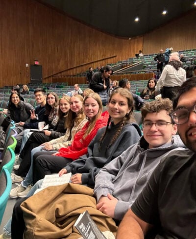 Selfie of nine young adults sitting on teal seats in a brown, high-roofed auditorium.