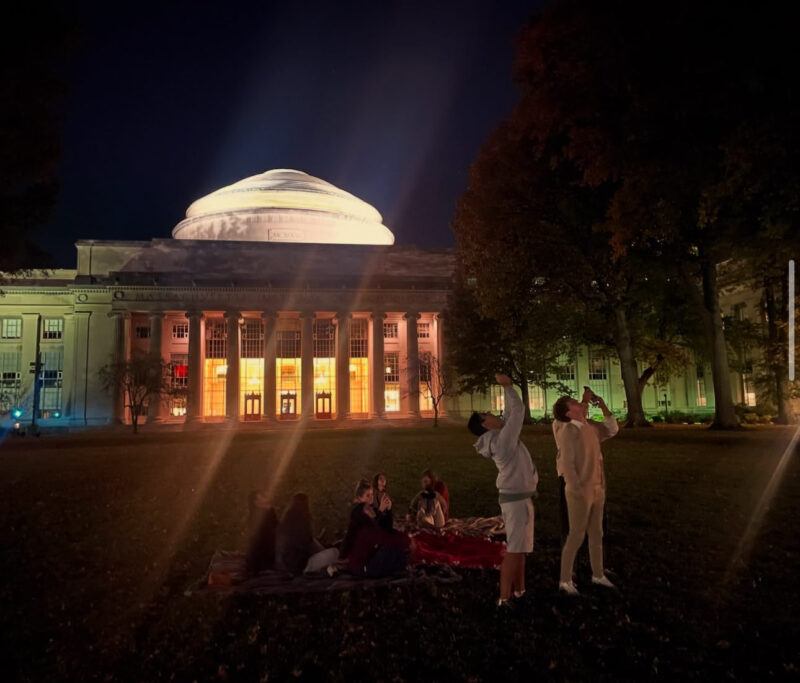 A group of teens on MIT's Killian Court at night, in front of the Great Dome. Some are sitting on a picnic blanket in the grass, others are standing up taking pictures of the sky. There is warm lighting coming from the building behind them