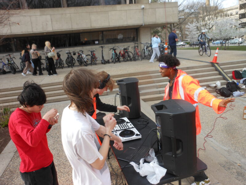 Three people stand behind a table, one of them DJing. A man in one of the orange safety jumpsuits talks to them excitedly
