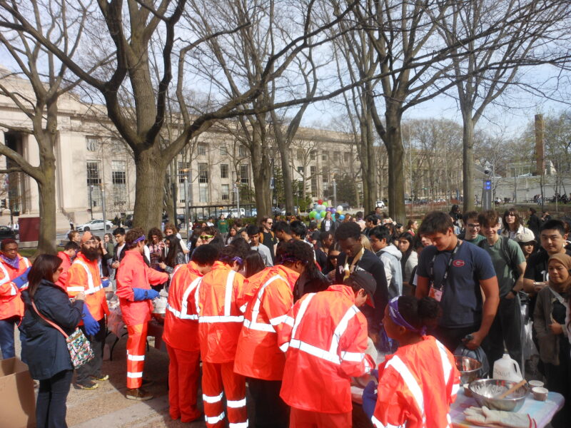 a group of people in bright orange jumpsuits stand at a table outdoors, mixing ice cream in bowls. Only their backs are visible, so you can see the large crowd that has gathered around the table to get ice cream. It's a sunny day, leaves are beginning to sprout on some of the trees surrounding them, and the main MIT building is visible in the background.