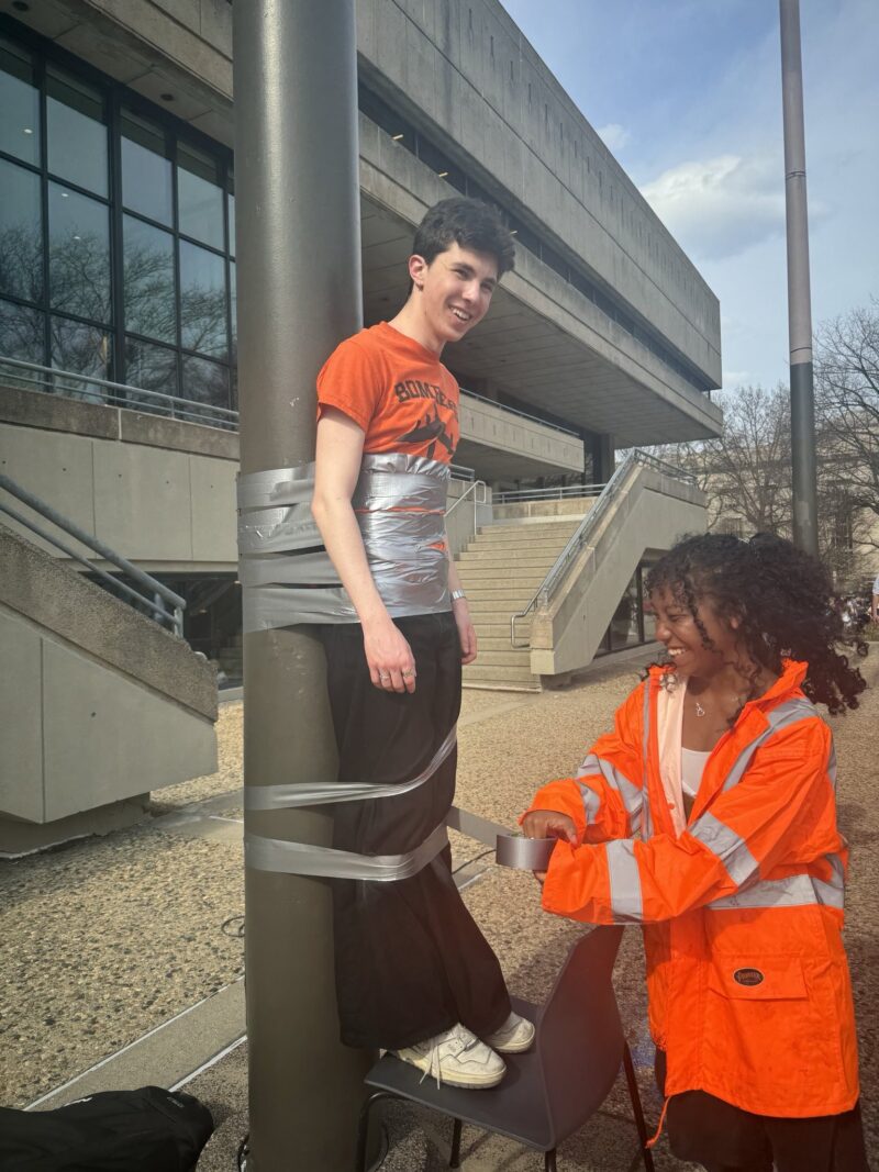 A boy in an orange shirt smiling as a girl in bright orange PPE duct tapes him to a pole