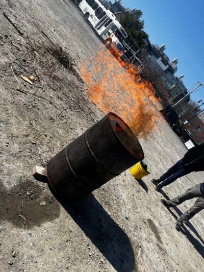 A large outdoor barrel burns intensely, with flames pushed horizontally by the wind. Twigs and brush lie on the ground nearby. Two people stand at a distance observing, with an MIT bus and campus buildings visible in the background.
