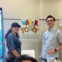 Three smiling individuals pose in front of a handmade sign that reads “Water Scope” at…