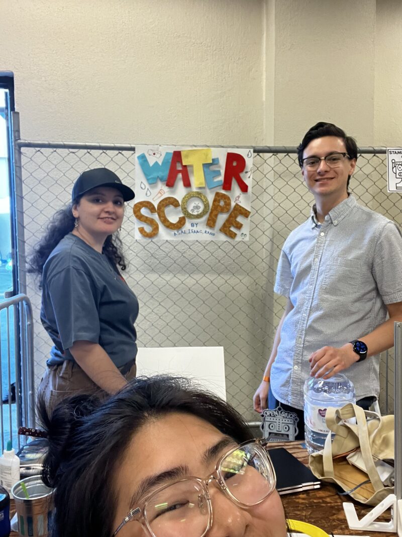Three smiling individuals pose in front of a handmade sign that reads “Water Scope” at a booth. The person on the left has long curly hair and wears a dark cap and blue shirt. The person on the right wears glasses and a light button-down shirt, holding a large water jug. In the foreground, another person with glasses and a joyful expression partially fills the frame. The setup includes art supplies and a gridded fence behind the display.
