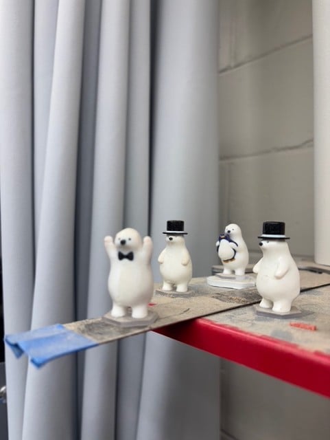 A scene on a dusty shelf. A few polar bears in tophats stand around an extending ruler. At the end of the ruler gangplank is a polarbear without a tophat, hands in the air. Another polarbear stands nearby, watching, clutching a small penguin. It looks like he's getting