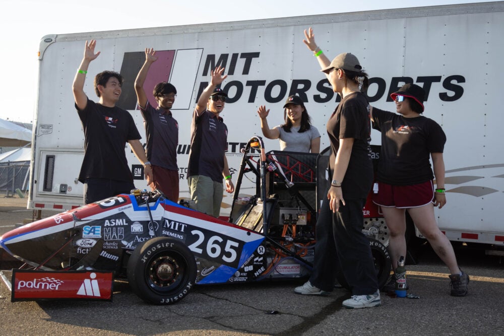 A group of people throwing their hands up, behind a red and black racecar