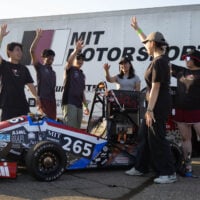A group of people throwing their hands up, behind a red and black racecar