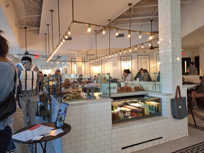 Modern bakery and café with white tiled walls, glass display cases of pastries and drinks, and customers waiting in line near the counter.