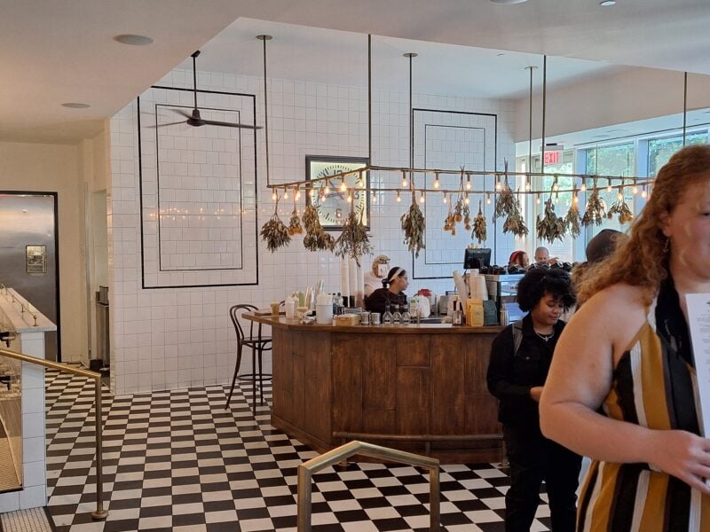 A café with checkered floor, a wood counter, and hanging dried herbs. People are ordering drinks, and large windows let in natural light.