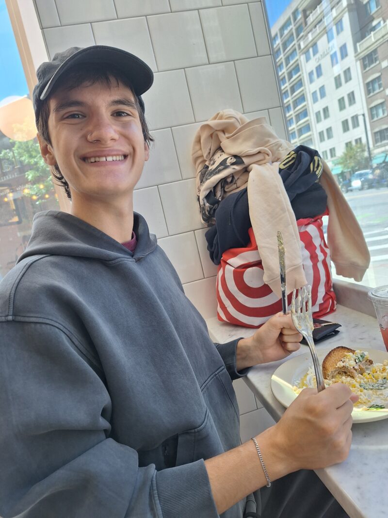 Smiling person in a hoodie and cap sits at a café with a plate of food. The scene is bright with urban buildings visible through the window. Warm and casual mood.