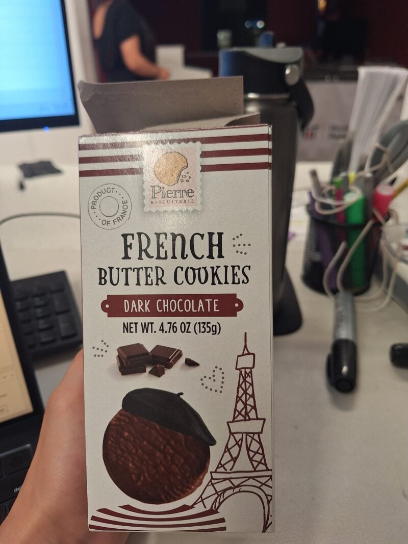 A hand holding a box of Pierre French Butter Cookies in Dark Chocolate, featuring an Eiffel Tower illustration and a snack-related design.