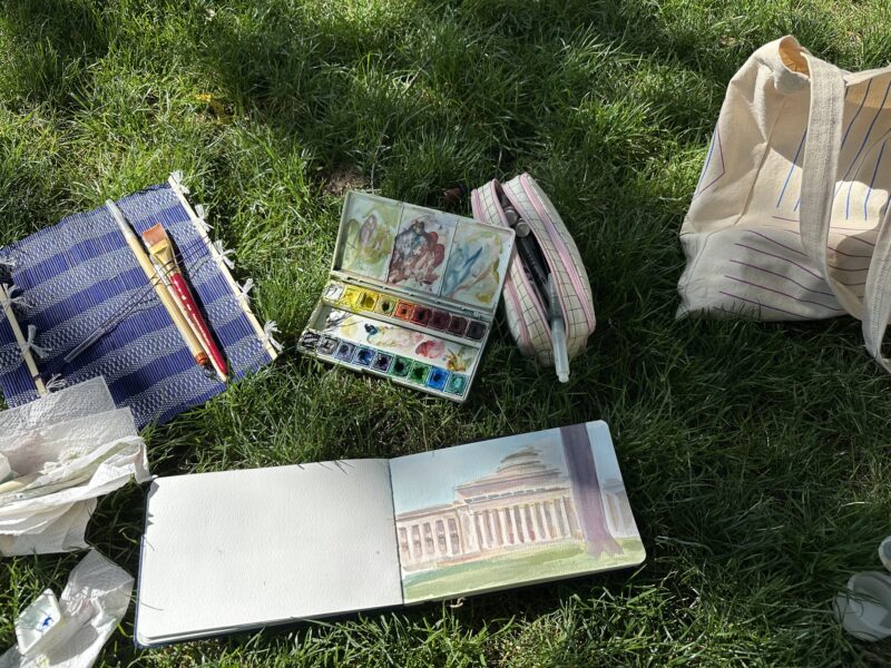 sketchbook, watercolors, paintbrushes, bag strewn on grass