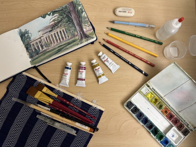 sketchbook, paint, brushes, paint palette, colored pencils, eraser, water, etc. strewn on table