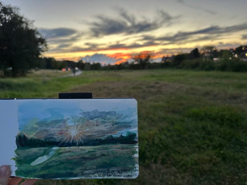 painting of a sunset over a field with same scene IRL in back