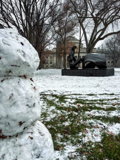 photo of snowman in close foreground, with partially snow-covered lawn and statue in background; building 10 looms in distance