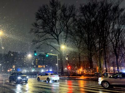 image of street at night full of cars that is wet with snowmelt; snow particles flutter in front of the traffic signals
