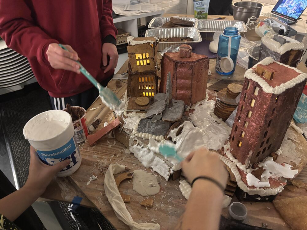 a progress shot of people putting icing and marshmallow fluff onto a gingerbread structure