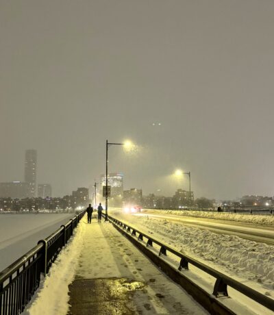 sidewalk of snow-covered bridge with frozen river on the left-hand side and melancholy streetlights in the distance
