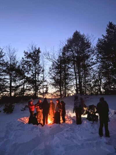 people huddling around a campfire surrounded by thick snow in the foreground with trees and sunset in the background