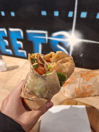 Hand holding a pita wrap filled with colorful vegetables, wrapped in brown paper. Background shows graffiti that reads "EETS." Casual street food vibe.