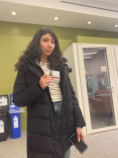 Person standing indoors wearing a long black winter coat and holding a phone while showing an ID card toward the camera. A green wall, recycling bins, and a small glass room are visible in the background.