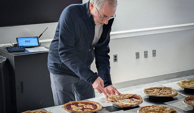 Stu stands in front of a table and is cutting into one of the pies.