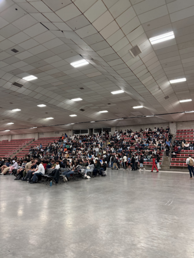 Wide shot of Johnson Ice Rink as attendees stream in for the CPW opening ceremony. Hundreds of people fill the gray folding chairs on the rink floor and the red bleacher seats along the far wall. The large concrete ceiling is lit by fluorescent panels. A figure in a bright red outfit walks along the side of the room.