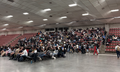 Wide shot of Johnson Ice Rink as attendees stream in for the CPW opening ceremony. Hundreds of people fill the gray folding chairs on the rink floor and the red bleacher seats along the far wall. The large concrete ceiling is lit by fluorescent panels. A figure in a bright red outfit walks along the side of the room.
