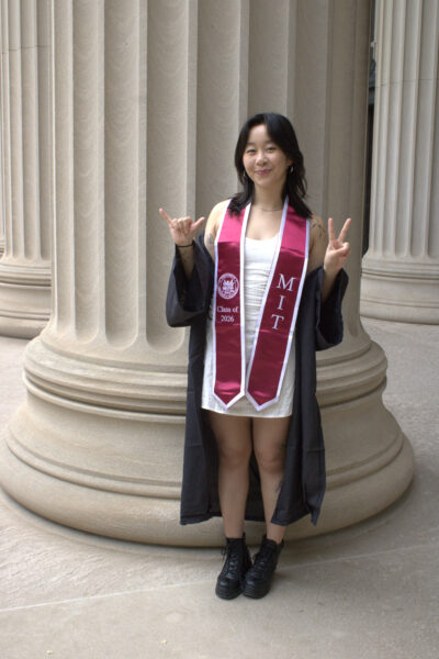 a girl poses in front of a pillar holding up a 6 and a 2, in graduation regalia