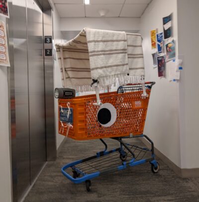 orange shopping cart in a hallway with large eyes on the side, a blanket canopy, a liscense plate, and a large flashlight