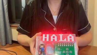 A person sitting at a table holds a colorful LED project display with red lights spelling “HALA.”
